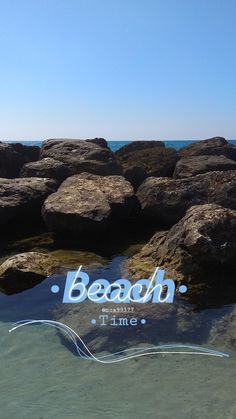 the beach time sign is reflected in the water near some rocks on the shore line