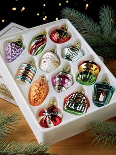 an assortment of christmas ornaments in a white box on a wooden table next to pine branches