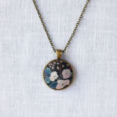 Inspired by Liberty Art Fabric, I developed my collection of vintage-style jewellery. The vibrant floral motifs of Liberty fabric are truly celebrated and showcased in this collection. A great gift for nature-lovers that will never go out of fashion. This necklace features white flowers in the dark background which has an oriental painting feeling. It has an antique bronze alloy pendant tray and an oval pendant. The long brass chain has a lobster clasp and a tiny bee beside it. Pendant 2.8 x 2.8 Vintage Flower Charm Necklace For Gift, Flower Shaped Necklace With Vintage Charm As Gift, Flower Shaped Necklace With Vintage Charm For Gift, Handmade Vintage Jewelry With Round Pendant, Multicolor Brass Elegant Necklaces, Elegant Multicolor Brass Necklaces, Vintage Necklaces With Flower Charm Round Pendant, Vintage Flower Charm Pendant Jewelry, Vintage Charm Flower Pendant For Jewelry Making