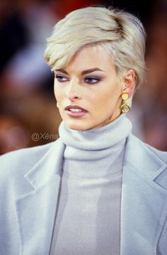 a woman with blonde hair and blue eyes wearing a gray coat, grey turtle neck sweater and gold earrings