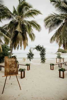 a sign that is sitting in the sand near some palm trees and chairs with writing on it