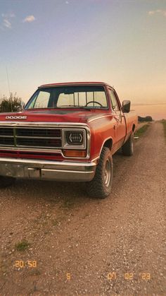 an old red truck parked on the side of a dirt road next to a field