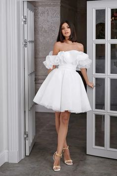 a woman standing in front of a door wearing a white dress with ruffled sleeves