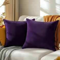 two purple and orange pillows on a white couch in front of a vase with flowers