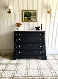 a black dresser with three drawers in front of a painting on the wall behind it