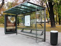 a bus stop sitting on the side of a road next to a trash can and tree