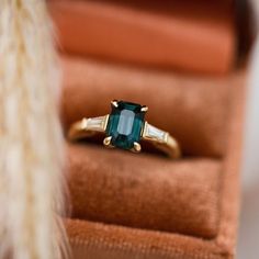 an emerald and diamond ring sitting on top of a velvet box next to a feather