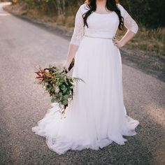 a woman in a white dress is standing on the side of the road holding a bouquet