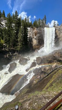 a waterfall with a rainbow in the middle and trees around it on a sunny day