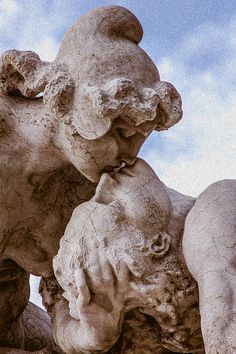 two stone statues with one holding the other's face to their mouth, in front of a blue sky and white clouds