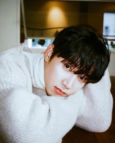 a young man with black hair wearing a white sweater and looking at the camera while sitting on a wooden floor