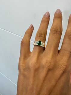 a woman's hand with an emerald and diamond ring on her left hand, against a white wall