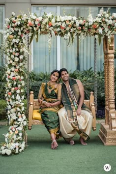 two people sitting on a swing in front of a flowered arch with white and orange flowers