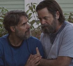 two men sitting next to each other in front of a house and one is holding his hands together