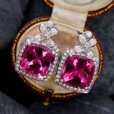 These collectable earrings are each accented with one (1) cushion cut natural Rubellite tourmaline set into a four-prong setting. The earrings are also each accented with thirty-two (32) bezel/ bead set round brilliant cut diamonds. The diamonds are bordered with milgrain edging. The earrings measure 19.5mm by 12.7mm and are finished with posts and friction backs for pierced ears. Cushion Cut Gemstone Earrings For Formal Occasions, Fine Jewelry Cushion Cut Gemstone Earrings, Cushion Cut Gemstone Earrings In Fine Jewelry Style, Cushion Cut Gemstone Earrings Fine Jewelry, Cushion Cut Gemstone Earrings For Fine Jewelry, Elegant Cushion Cut Gemstone Earrings, Elegant Tourmaline Gemstones With Accents, Luxury Tourmaline Earrings As Gift, Luxury Tourmaline Earrings For Gift