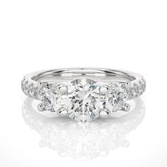 "This stunning brilliant trellis three stone engagement ring is a masterpiece of fine craftsmanship and exquisite design, perfectly combining the beauty of a 1.20+ carat Round lab-grown diamond with the sparkle of three stone diamonds. The Round diamond is of exceptional quality, with brilliant cut facets that maximize its natural radiance and fire. The diamond is set in a three stone shank design, which adds depth and dimension to the ring by framing the center stones appear to float invisibly above the surface of the ring. The ring's shank is also set with pave diamonds, which create a glittering band of brilliance that draws the eye toward the center stone. Each diamond is expertly set in a shared prong setting, which ensures that each stone is securely held in place while allowing maxi Luxury White Three-stone Moissanite Wedding Ring, Luxury White Moissanite Three Stone Wedding Ring, Three Stone Lab Grown Diamond Promise Ring, Anniversary Three Stone Moissanite Jewelry, White Moissanite Three-stone Ring, Three Stone Moissanite Jewelry For Anniversary, Lab Grown Diamond Three Stone Promise Ring, White Three Stone Moissanite Rings, White Moissanite Three Stone Rings