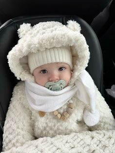 a baby in a car seat wearing a white hat and scarf with a pacifier