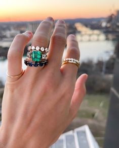 a woman's hand holding an emerald and diamond ring with the sun setting in the background