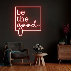 a neon sign that says be the good in front of a black wall and wooden floor