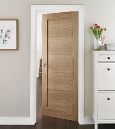 an open door in a white room next to a chest of drawers and a vase with flowers