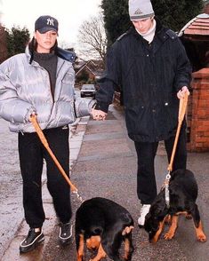 two people are walking their dogs down the street on leashes and one is wearing a hat