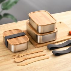 three stainless steel containers with bamboo lids and spoons on a cutting board next to utensils