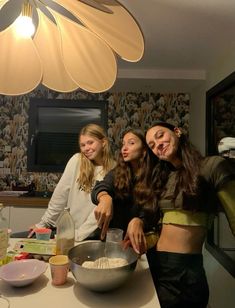 three girls in the kitchen mixing ingredients for some kind of cake mix, and one girl is holding a whisk with her right hand