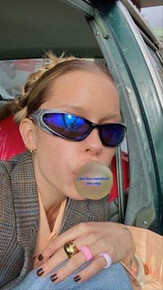 a woman wearing sunglasses and holding a bubble in her mouth while sitting in the back seat of a car