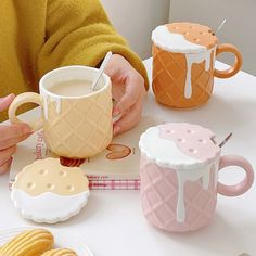 a person holding a cup and some cookies on a table with two mugs next to each other