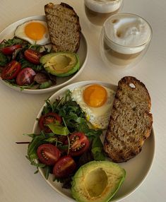 two white plates topped with eggs, bread and salad next to cups of coffee on a table