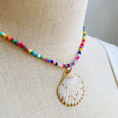 Embrace the essence of summer with this exuberant, colorful beaded necklace, featuring a stunning natural seashell pendant encased in a delicate golden trim. Each bead is meticulously selected to create a kaleidoscope of joy around your neck, reminiscent of a sun-kissed day at the beach or a vibrant festival dance. The heart of this necklace is the seashell pendant, a treasure from the ocean's depths, polished to perfection, and framed in gold. This piece is a celebration of nature's artistry, w Bohemian Shell-shaped Jewelry With Colorful Beads, Beach Shell Jewelry With Tiny Beads, Colorful Shell Bead Necklaces For Vacation, Beaded Shell-shaped Jewelry For The Beach, Colorful Beads Shell Necklace For Vacation, Shell-shaped Beaded Jewelry For Beach Season, Bohemian Beaded Shell-shaped Necklaces, Bohemian Shell With Colorful Beads For Gift, Multicolor Strand Jewelry For Beach Season