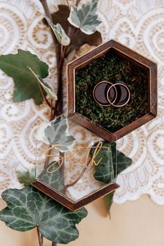 two wedding rings are placed in a wooden box surrounded by greenery and leaves on a lace doily