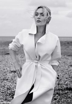 a woman is standing on the beach in a white coat and black pants with her hands on her hips