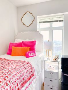 a white bed with pink and orange pillows in a bedroom next to a window,