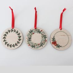 three wooden ornaments hanging from red ribbon on white background, one with holly wreath and the other with berries