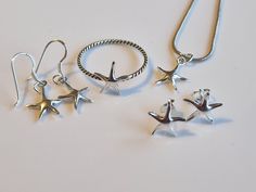 Discover the enchantment of the ocean with this exquisite starfish jewelry set, crafted to capture the wonder of the sea. This set includes a delicate necklace, a stylish ring, a pair of elegant stud earrings, and a pair of dangling earrings, all featuring the iconic starfish design. Each piece is finely crafted from high-quality silver, showcasing the intricate details of the starfish's arms and texture. The necklace's pendant gracefully hangs from a sleek silver chain, while the ring's twisted Starfish Design, Starfish Jewelry, Jewelry Ocean, Twisted Band, Stylish Rings, Dangling Earrings, Delicate Necklace, Beach Jewelry, Necklace Earrings