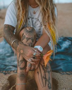 a woman sitting in the sand with tattoos on her arms and legs, holding an orange flower