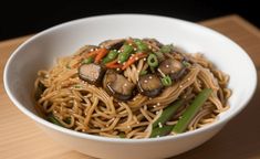 a white bowl filled with noodles, mushrooms and veggies on top of a wooden table