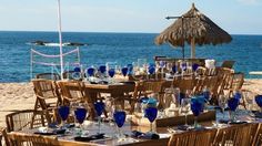 an outdoor dining area set up on the beach with blue glassware and straw umbrellas