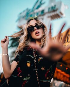 a woman wearing sunglasses standing in front of a neon sign with her hands out to the side