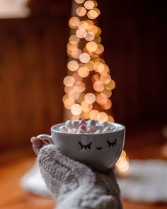 someone is holding a cup with marshmallows in front of a christmas tree