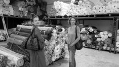 two women standing next to each other in a room filled with lots of rolls of fabric