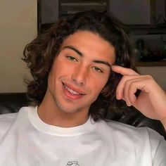 a young man with long hair is smiling and holding his hand to his ear while sitting on a couch