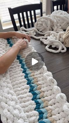 two people are crocheting together on a table with chairs and rugs in the background