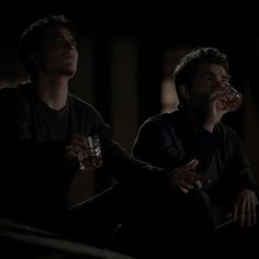 two men are sitting in the dark drinking from their glasses and looking up at something