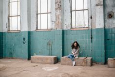 a woman sitting on concrete blocks in an old building with blue walls and windows behind her