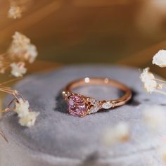 a pink diamond ring sitting on top of a gray surface next to dried white flowers