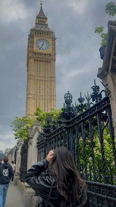 a woman standing in front of a fence with a clock tower in the back ground