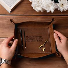 a person holding a brown leather tray with writing on it and some flowers in the background