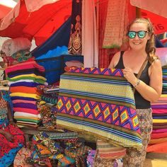 a woman standing in front of a display of colorful bags and blankets at an outdoor market